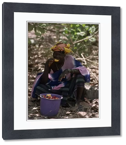 20075207. GAMBIA People Women Woman sat with bucket eating cashew fruit