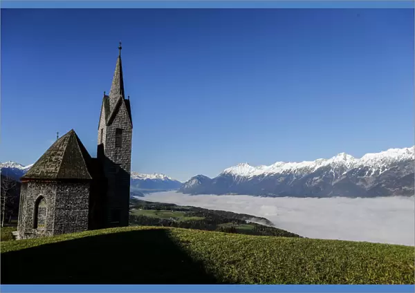 Church overlooks fog covered Inntal valley in front of snow covered mountain summits on a