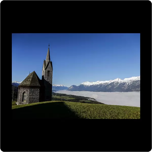 Church overlooks fog covered Inntal valley in front of snow covered mountain summits on a