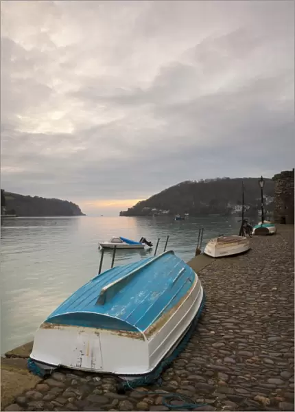 Small dinghies laying upside down on cobbled quay, with overcast sky at sunrise, Dartmouth Harbour, Dartmouth, Devon