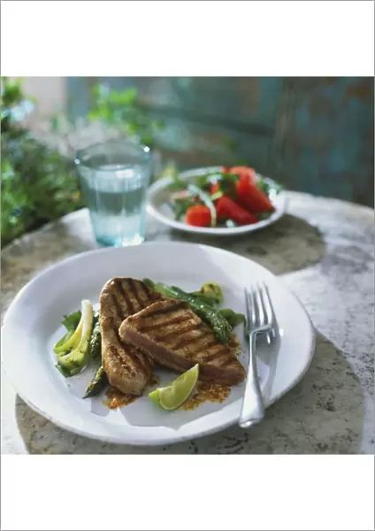 Chargrilled steaks of tuna with asparagus sticks, spring onion and lime slice on plate with fork, bowl of salad and glass of water in background, front view