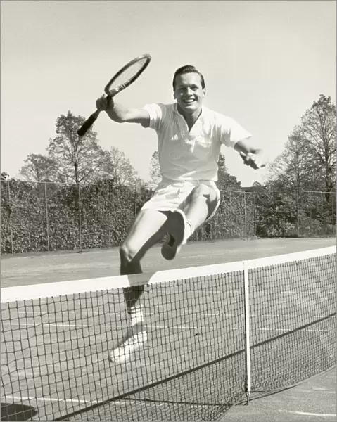 Man holding tennis racquet leaping over tennis net, 1950-60s, black and white