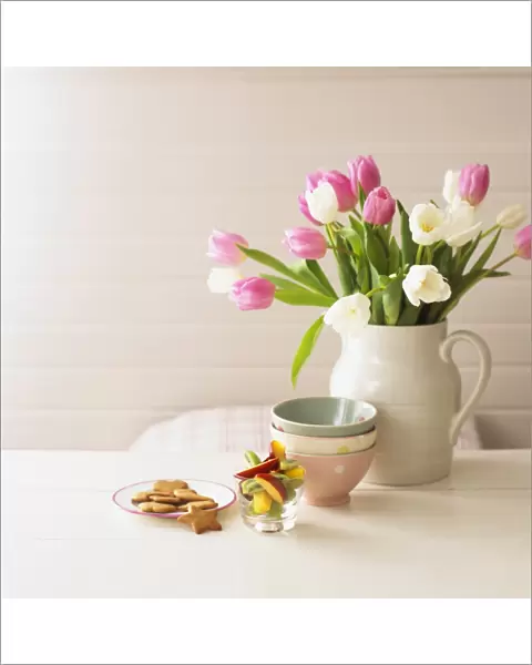 Jug containing tulips, stacked bowls, glass of sliced fruit, and plate of biscuits, all on table