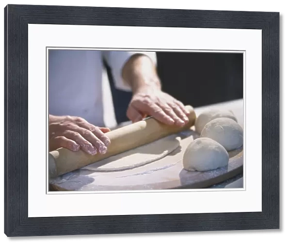 Three dough balls resting on large wooden board, thin layer of dough being rolled out with wooden rolling pin