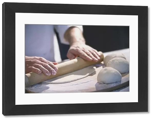 Three dough balls resting on large wooden board, thin layer of dough being rolled out with wooden rolling pin