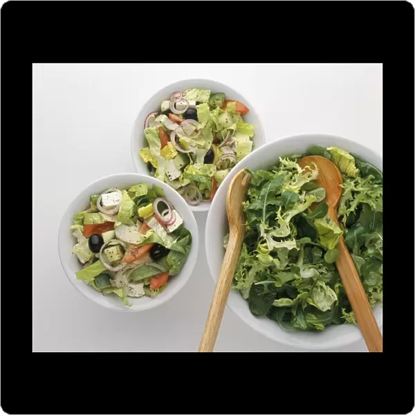 Two small bowls of Greek salad and large bowl of salad greens and wooden serving spoon and fork