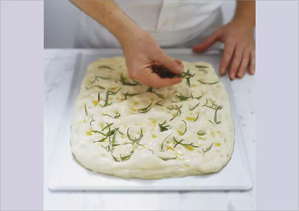 Fresh rosemary sprigs being pressed into uncooked focaccia dough, high angle view