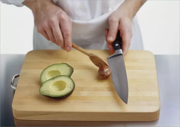 Using wooden spoon to pry avocado stone off knife blade, two avocado halves on chopping board