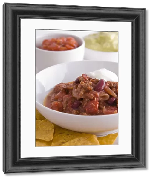 Bowl of chilli con carne with tortillas chips, soured cream and bowls of salsa and guacamole in background