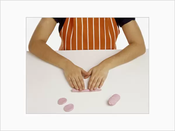 Hand model wearing an orange and white striped apron, making oval shapes from pink fondant icing, rolling icing into long bar