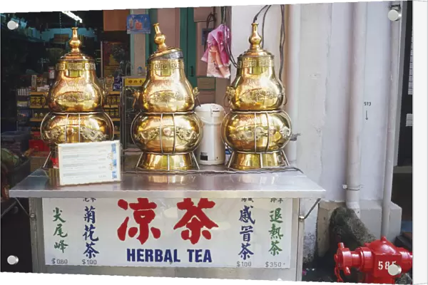 Singapore, Chinatown, Temple Street, decorative herbal tea dispensers, gold coloured metal decorated with Chinese script and dragons, lid loosely placed on central dispenser, standing on metal box, shop in background