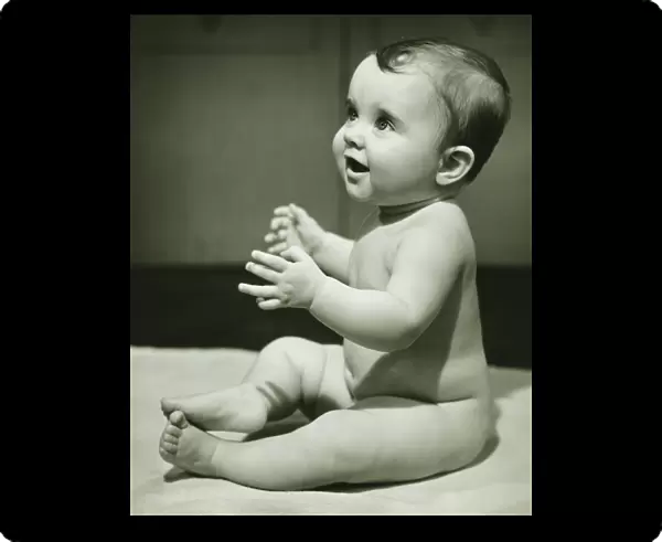 Baby boy (12-18 months) with raised hands, sitting on bedspread, (B&W), close-up