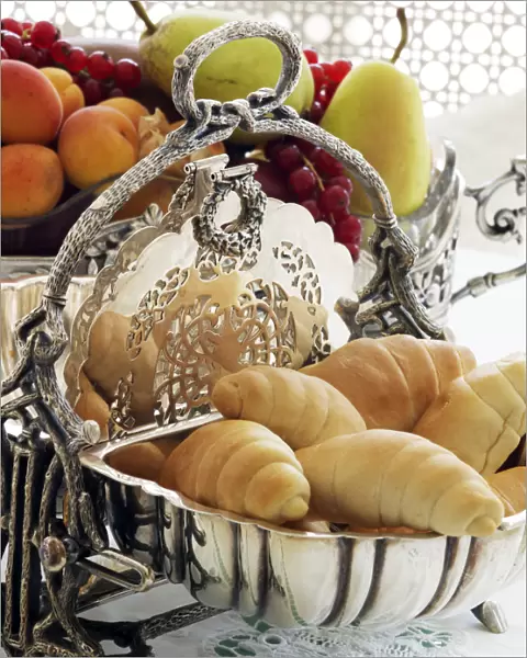 Antique silver hinged bread basket with croissants in front of a bowl of fruit on a breakfast table