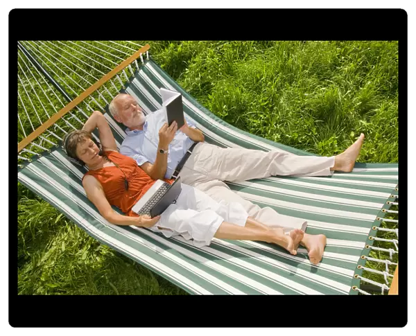 Senior citizen couple lying in a hammock, woman wearing a headset looking at a netbook or laptop, man reading a book