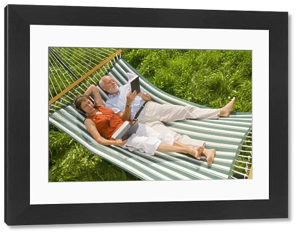Senior citizen couple lying in a hammock, woman wearing a headset looking at a netbook or laptop, man reading a book