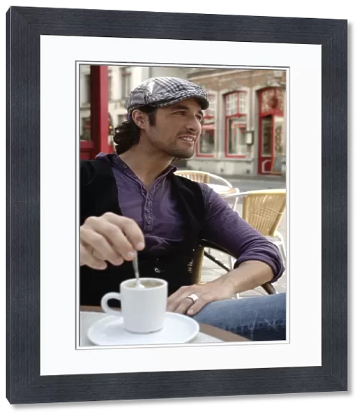 Man, early 30s, wearing casual clothes sitting in an outdoor cafAzA stirring a coffee cup, smiling