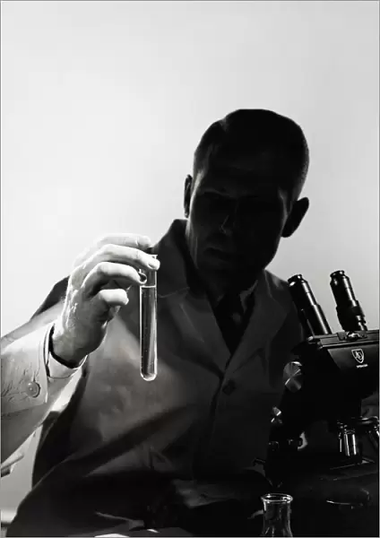 Male scientist wearing lab coat, sitting in darkness behind microscope