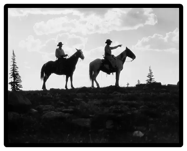 Silhouette of two cowboys on horses on top of mountain, one man pointing the way