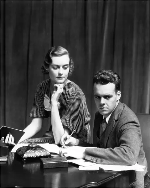 Couple Working On Budget At Desk Woman Sitting On Arm Of Chair Man With Pencil Writing On