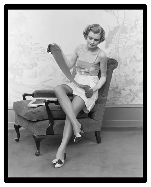 Woman wearing lingerie, sitting on arm of chair, holding up pair of silk stockings
