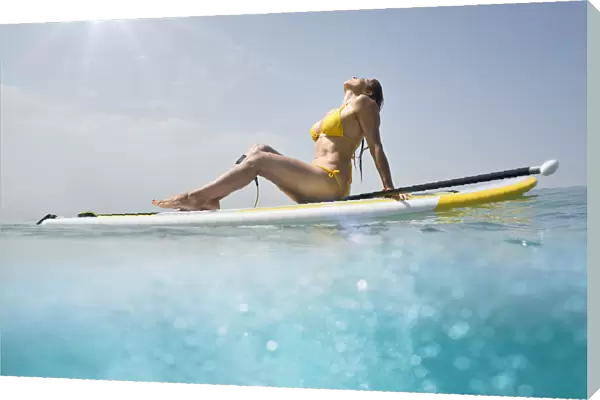 A woman in a yellow bikini sits with her paddle and board basking in the sunlight on the ocean; Tarifa cadiz andalusia spain