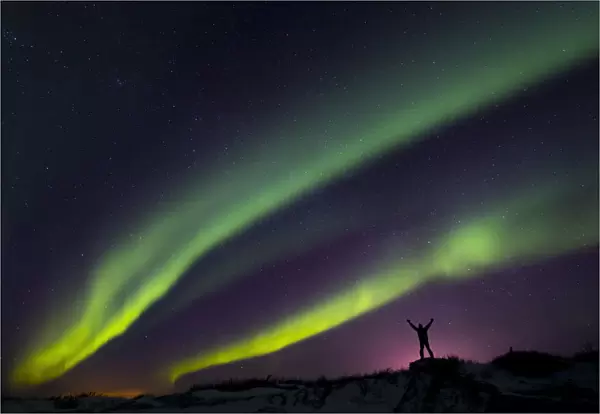 Colourful Aurora Borealis Over A Man With Arms Outstretched Silhouetted Against Light Pollution From Nearby Fort Greely; Alaska, United States Of America