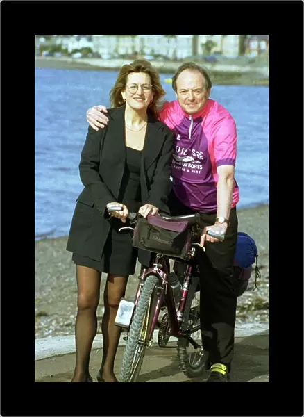 Barbara Dickson wearing glasses and a black suit poses with James Bolam on a bicycle in