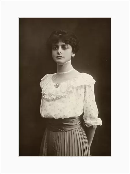 Three-quarter-length portrait of young woman standing, with her hands behind her back. She is wearing a pleated skirt and a ruffled blouse