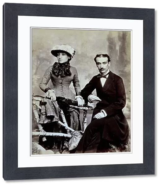 Three-quarter length portrait of a young couple. The man is seated and resting one arm on a fence. The woman is standing next to him