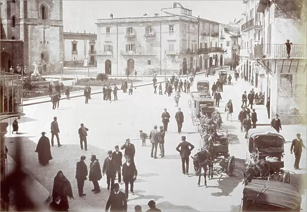 View of a square in c City in Sardinia. On the left side of the square, turned into a garden, is a clock tower. On the right a few horse-drawn carriages the square is filled with people