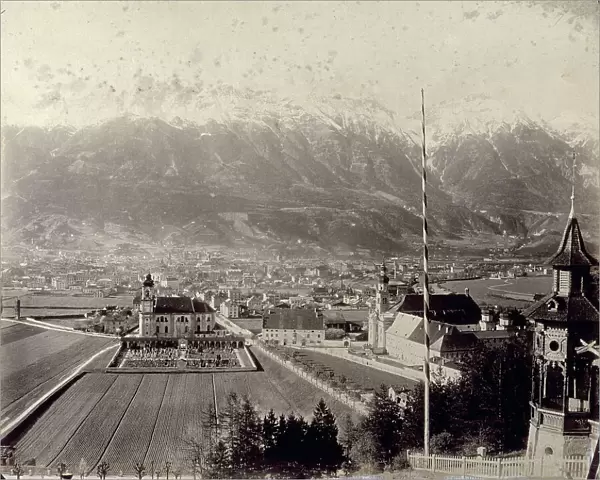 View of Innsbruck from the small hill of Berg Isel':in the foreground the Wilten Basilika flanked by a small cemetery and surrounded by ploughed fields; on the right is the Stiftskirche Sankt Laurentius'; the vast urban center stretches out in the background. In the far distance, the high mountains partially covered with snow
