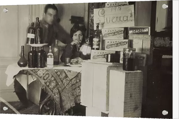 Portrait of a couple sitting at a table with bottles, boxes and sign with inscription '1918 War!'