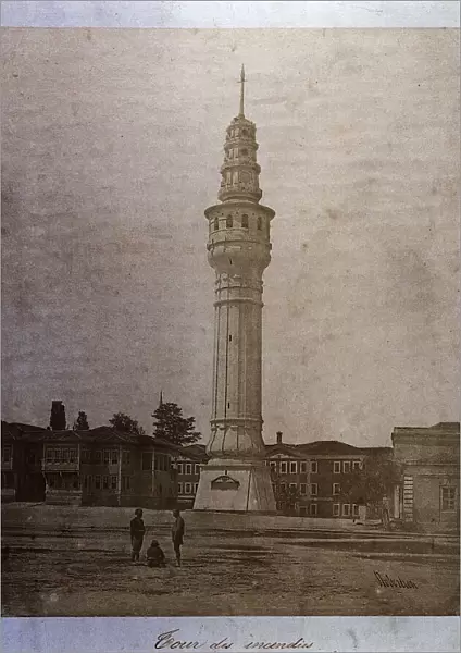 The Fire Tower in Istanbul (Constantinople) rises at the center of an open space among residential buildings. There are three people in the street in front of the tower