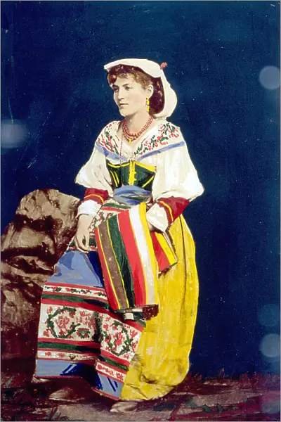 Full-length portrait of a young woman in a brightly colored traditional roman costume, with a coral necklace. She is posing in the studio, seated on a rock. She has a colored shawl on her left arm