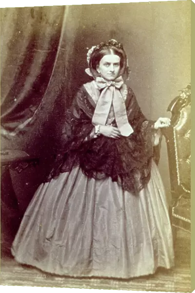Full-length portrait of an elegant lady in romantic period day dress, with a large lace shawl. She wears a hat tied nder her chin with a large bow