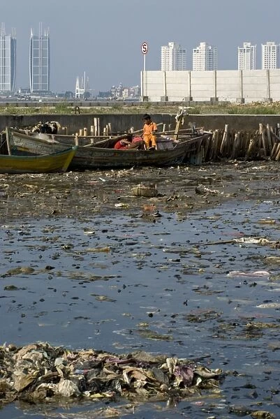 Children on boat in rubbish filled water, with city skyscrapers in background, Muara Karang, Jakarta, Java, Indonesia