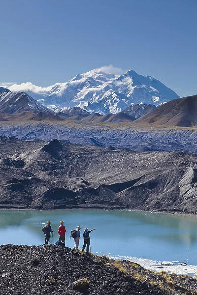 A group of hikers stand on moraine next to a glacial meltpond at the edge of the Muldrow Glacier