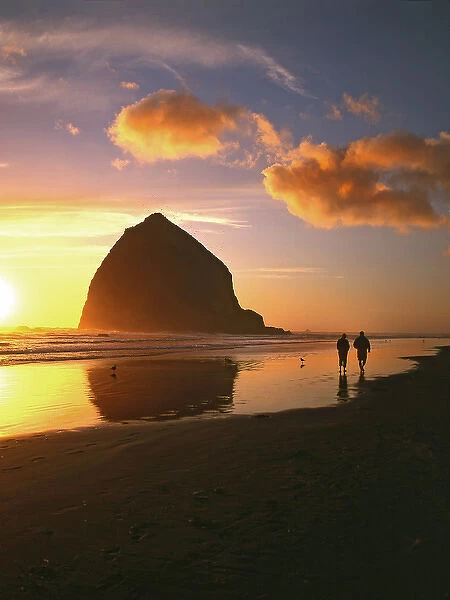 USA, Oregon, Cannon Beach. Couple walking beside Haystack Rock at sunset. Credit as