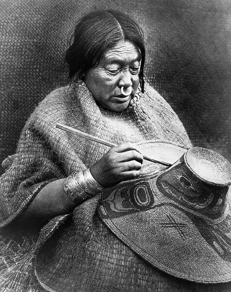 The daughter of a Nakoaktok chief on the coast of British Columbia, Canada, wearing a cedar bark cape and a nose-ring made of abalone shell, and painting a crest on a hat made of woven shreds of spruce roots. Photographed by Edward S. Curtis, 1914