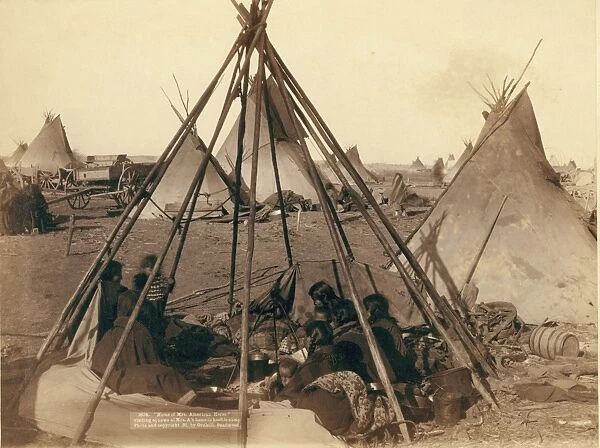 Oglala women and children seated inside an uncovered tepee frame in an encampment