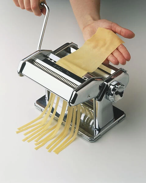 Thin sheet of pasta dough being rolled through pasta machine and cut into strands, high angle view