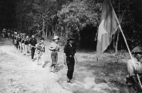 South vietnamese guerrillas file through the jungle along the ho chi minh trail, the flag with a yellow star on a red and blue background is always carried into battle, vietnam war, mid 1960s