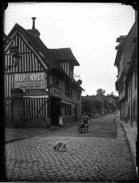 France, Basse-Normandie, Calvados (14), Pont-l'Eveque (Pont l'Eveque): rue bordered by medieval half-timbered houses, 1900 - Advertisements: Dubonnet, wine tonic at the quinquina grand prix 1900, continental tire with strong canvases