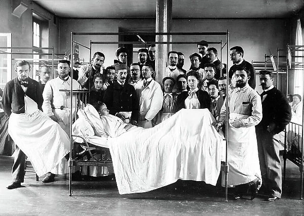 France, Ile-de-France, Paris (75): hospital, a boss and his whole team in professional pose: supervisor, nurses, interns around an old sick woman curled in bed - Reflex hammer and stethoscope, 1890