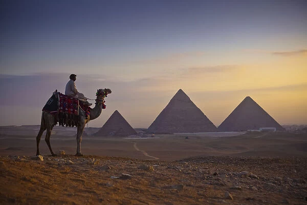 A Lone Camel And Rider Stand In Front Of The Setting Sun With The Great Pyramids Behind Them; Giza, Egypt