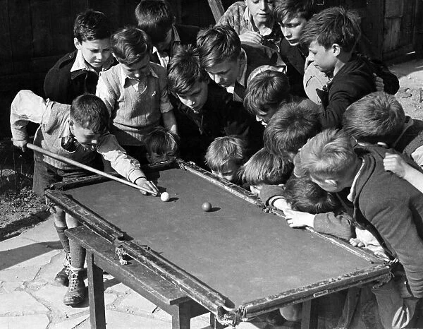 Children gather around a snooker table to play. Sixteen tousled heads bend over a rickety