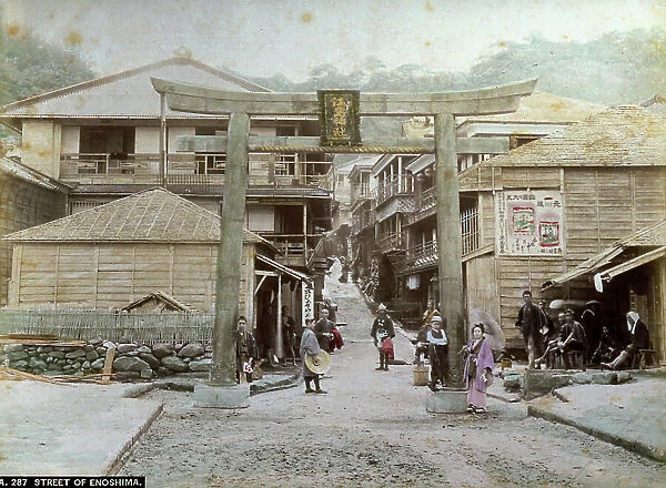 A busy street in Enoshima, in Japan, flanked by humble buildings. Isolated in the foreground is a typical Torii gate