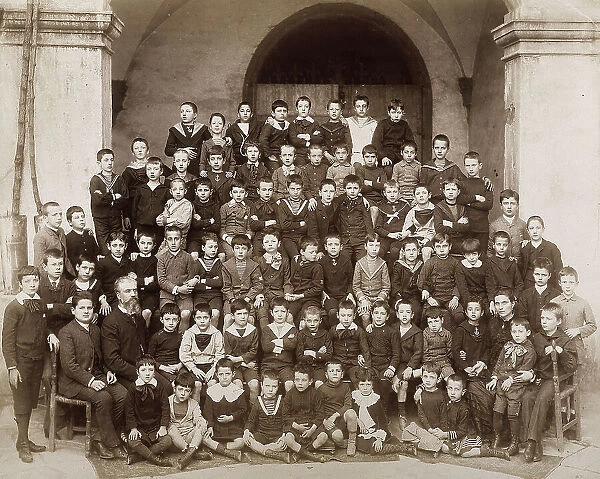 Large group of children, posing in rows in a pyramid form. They are probably in the courtyard of a public school or a boarding school and are almost all wearing sailor outfits. The teacher is seated in their midst