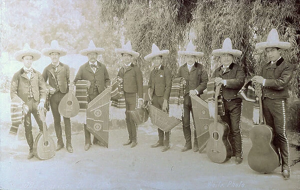 Members of a Mexican band pose in a garden. Standing next to each other, they wear identical costumes and sombreros. Each of them has a poncho on one arm and holds his instrument in the other