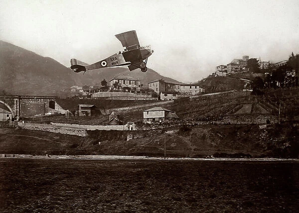 An S.V.A. warplane flies over a mountain village, in the background a bridge is visible